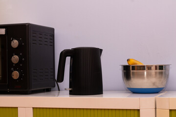 Black electric kettle and stainless steel bowl with banana on countertop beside microwave, showcasing modern kitchen appliances and vibrant colors in a cozy home environment