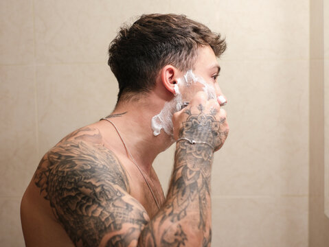 Young man with tattooed arms shaving in the bathroom. He applies shaving foam to his face and uses a razor while looking into the mirror, capturing a personal grooming and self-care moment