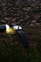 Smartwatch resting on a moss-covered log, illuminated by a flashlight, showcasing modern technology in a natural outdoor setting with autumn leaves scattered around