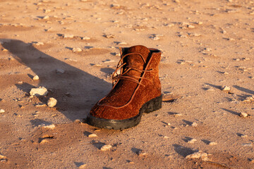 Brown suede boot resting on sandy terrain, surrounded by small rocks, showcasing rugged outdoor footwear in a natural setting, ideal for adventure enthusiasts and fashion lovers