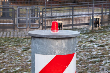Red plastic cups stacked on a gray metal barrier with a white and red striped design, surrounded by grass and a blurred background of a fenced area