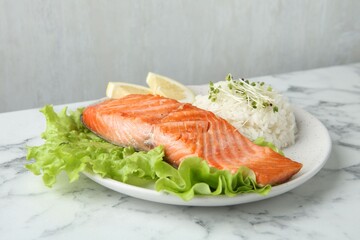 Piece of tasty grilled salmon, lemon, lettuce, rice and microgreens on light marble table against grey background, closeup