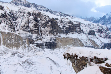 Mountain peaks covered in snow are seen in the distance. A large snow-covered rock is in the foreground, complete with icicles. Location is the Annapurna Base Camp in Nepal