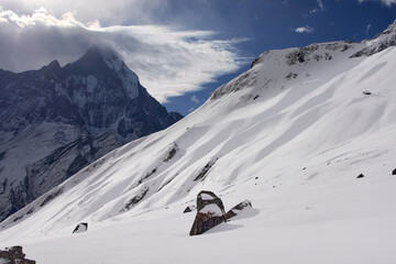 A snowy mountainside is visible, with a large peak in the distance partially obscured by clouds. The scene takes place within the Annapurna Sanctuary, Nepal. It is daytime.