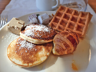 A generous breakfast plate featuring fluffy pancakes dusted with powdered sugar, a glazed waffle, buttery croissant, and assorted pastries arranged on a white dish with a coffee cup in the background.