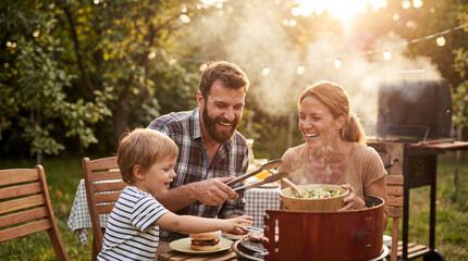 Happy Caucasian family having fun during a backyard barbecue, with a laughing father grilling burgers and a mother serving salad to their young son at a wooden table.