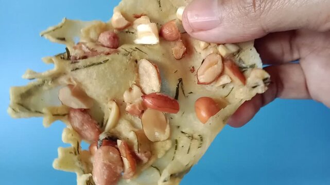 Hand showing the detailed texture of an Indonesian peanut cracker (rempeyek kacang), highlighting toasted peanuts and sliced kaffir lime leaves.