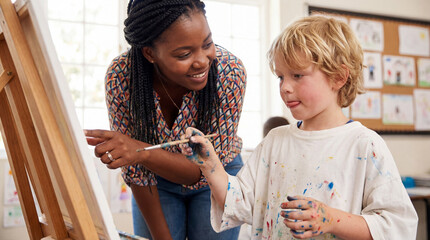 Happy young Black female teacher guiding a little blonde boy with messy hands as he paints on an easel during an art lesson in a bright elementary school classroom.