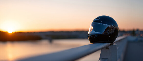 Black motorcycle helmet resting on white railing with blurred sunset and river background in soft warm light