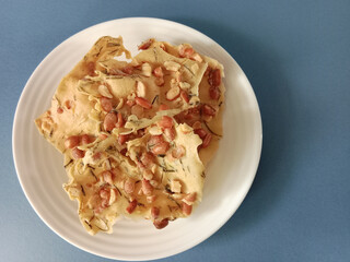 Crisp Indonesian peanut crackers (rempeyek kacang) piled on a white plate, captured from top view on a clean blue background with copy space for branding or advertising use.