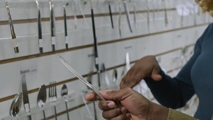 Close-up of hand of anonymous African American man holding and trying out sample table knife from display while shopping together for cutlery with wife in kitchenware store at weekend