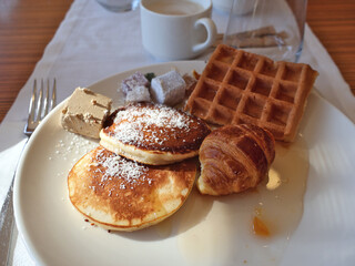A hearty breakfast plate featuring fluffy pancakes dusted with powdered sugar, a croissant, a waffle, butter, and coffee for a comforting morning meal.