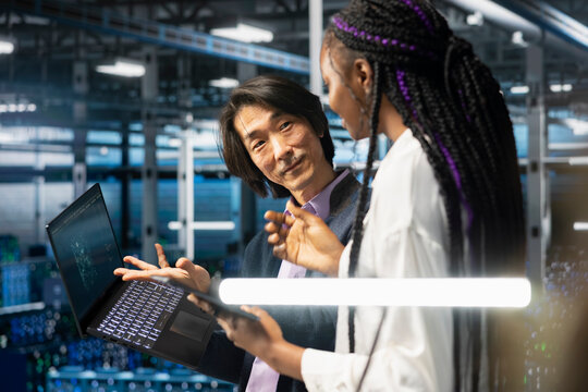 Multiethnic coworkers in diverse data center installing upgraded server racks with AI. African american woman and Asian senior programmer in server room upgrading gear using artificial intelligence