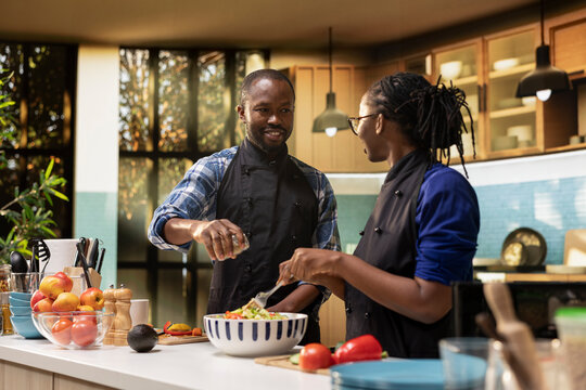 Black boyfriend adding spices and condiments in a salad from a jar, seasoning the organic lunch meal with fresh bio ingredients. Girlfriend mixing vegetables together in a bowl, vegetarian food.