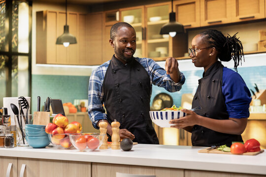 Happy couple preparing a fresh salad together in a bright kitchen, man adding a pinch of salt and the woman mixes colorful vegetables in a bowl. Healthy lifestyle of vegetarian people.