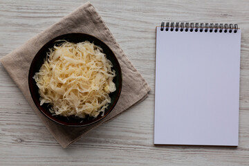 Organic White German Sauerkraut in a Bowl, blank notepad, top view. Overhead, flat lay.