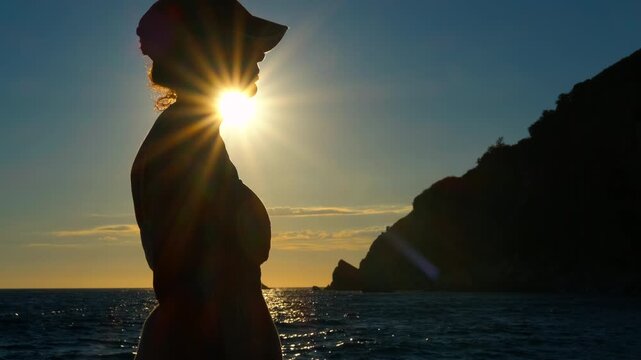 Silhouette of a woman kissing the sun at sunset over the sea. Side profile silhouette of a woman in a cap standing on a boat and looking at the ocean during a golden sunset, with sun flares