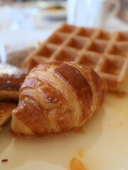Freshly baked croissant sits on a white plate with a soft-focus waffle in the background, creating a cozy breakfast scene. Perfect for bakery, cafe, morning meals, and French pastry themes.