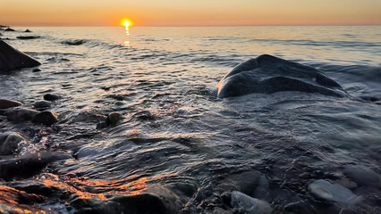 Calm waves roll over a rocky shoreline at sunset, with the golden sun reflecting on the tranquil sea surface.