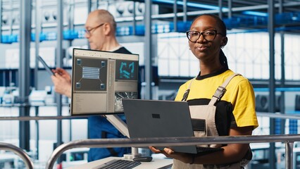 Smiling engineer overseeing smart factory, looking at collected metrics on laptop screen. Happy technician on platform in industry 4.0 industrial plant equipped with sensors using notebook, camera A