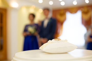 A wedding ceremony. In the foreground is a white ring pillow with two wedding rings on it. In the...