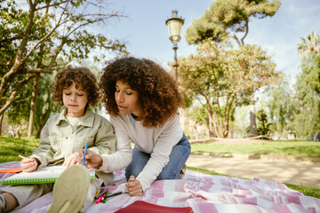 A mother and her son are drawing in a sketchbook and sitting on a blanket while she holds a felt-tip pen and he holds a pencil