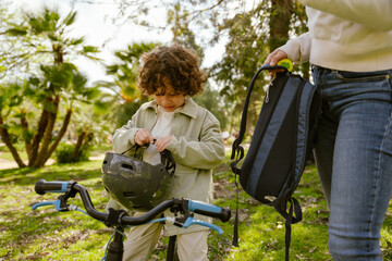 Fototapeta premium A son sits on a bicycle and unfastens his helmet while his mother stands next to him holding a backpack