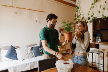 A man and a woman stand at a table while laughing and looking at a potted plant he is holding