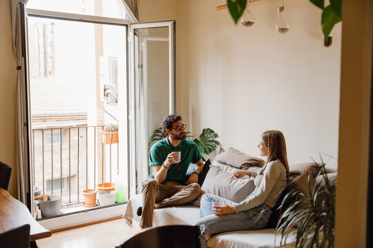 Man listening to smiling woman while holding glasses and sitting on sofa - Powered by Adobe