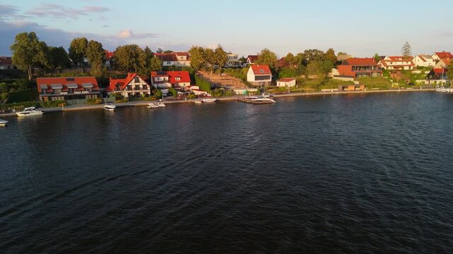 A picturesque summer evening in Mikolajki, Masuria showcasing colorful houses along the lakeside. Boats gently bob on the water while trees sway in the light breeze.