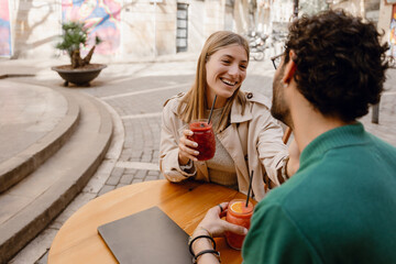 Woman laughing and holding hand next to man while they hold cups and sit at table