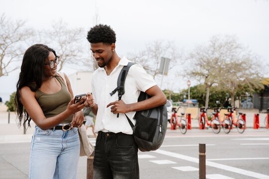 A man looks at a phone that a woman standing next to him is showing while they smile - Powered by Adobe
