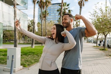A male athlete smiles and puts his hand on the shoulder of a female athlete who laughs as they flex their muscles and pose in front of a phone she is holding