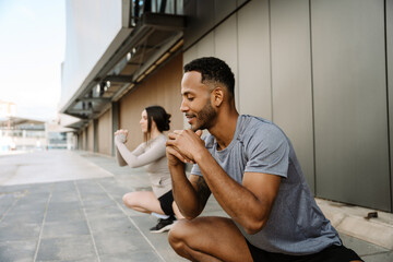 A male athlete smiles while squatting next to a female athlete, and they stretch their legs and...