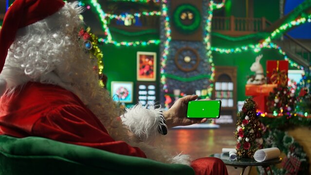 Santa Claus reads letters from kids on green screen mobile phone in Christmas ornaments adorn workshop. Old costumed man in holiday season ambiance holding mockup smartphone, camera A - Powered by Adobe
