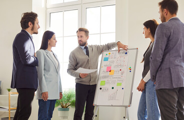 Confident businessman presenting strategy on whiteboard to diverse team in bright modern office. Professional team listening and discussing ideas during business meeting or brainstorming session.