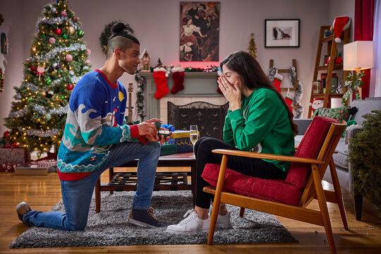 Man proposes to surprised woman with ring in festive Christmas living room decorated with tree and stockings