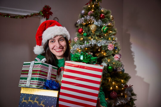 Woman wearing Santa hat holds wrapped Christmas presents in front of decorated tree with lights