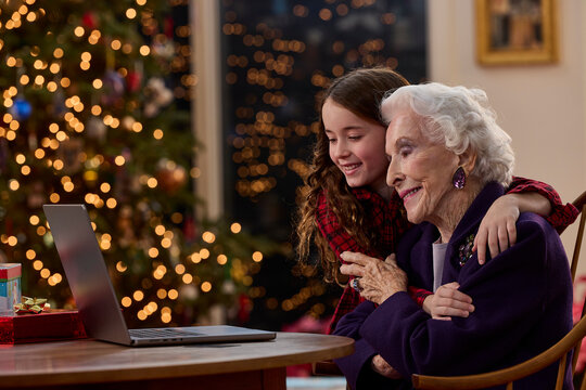Grandmother and granddaughter video calling family together during Christmas with festive tree lights in background