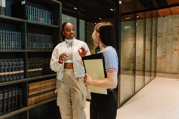 Female student gesturing and talking to female student standing next to her near bookshelves and holding folders