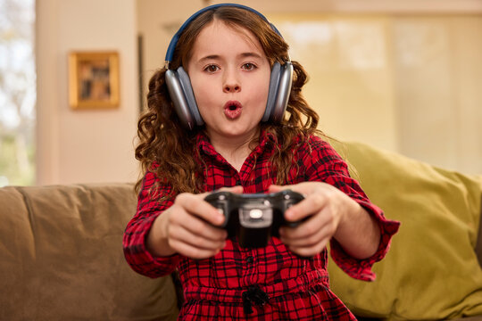 Young girl wearing headphones plays video games with controller on living room couch