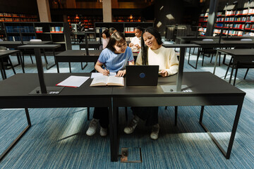 Two female students talking and looking at a laptop while sitting at a table and smiling while one of them holds a pen over a notebook