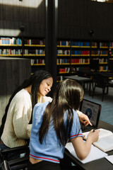 Female student smiling and pointing at a book over which a female student is holding a pen while they sit on chairs at a table in front of a laptop