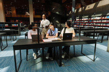 A group of three female students sit at a table while one of them talks to a female student walking next to them while they smile