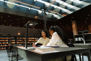 Female student holding pen and notebook and smiling while listening to female student while they are sitting at table