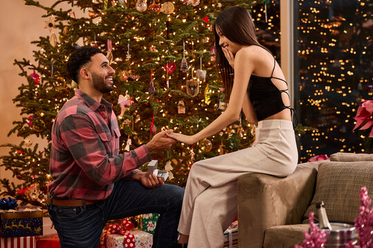 Man proposing to surprised woman with engagement ring in front of decorated Christmas tree