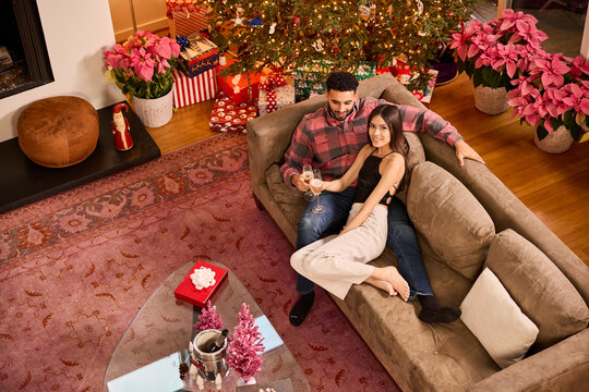 Couple enjoying champagne together on cozy sofa surrounded by festive Christmas tree and pink poinsettias