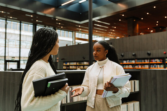Female student holding notebooks and standing near bookshelves while talking to female student holding laptop