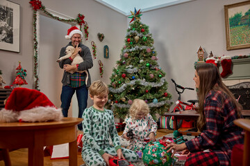 Family celebrating Christmas morning together with children opening presents and puppy by decorated tree