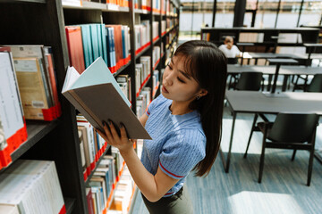 A female student reads a book she opens while standing near bookshelves